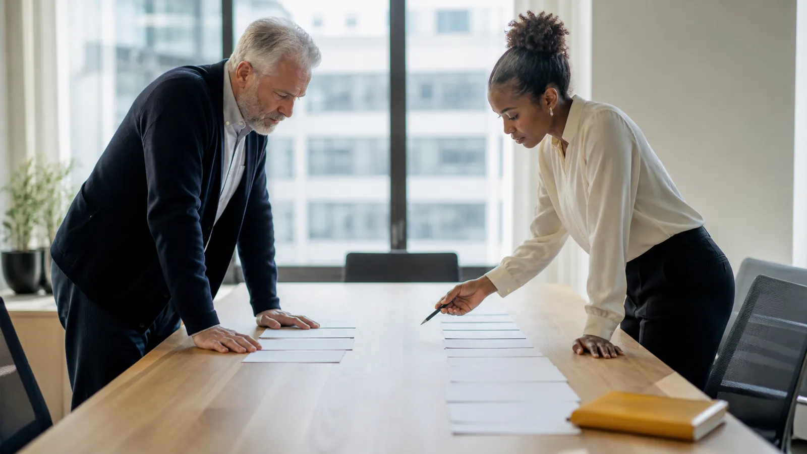 Two colleagues sorting worksheet pages into two columns on a conference table, comparing hiring paths